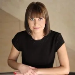 Portret kobiety w czarnej sukience, opartej o metalową barierkę, z delikatnym uśmiechem i profesjonalnym wyrazem twarzy, na neutralnym tle.