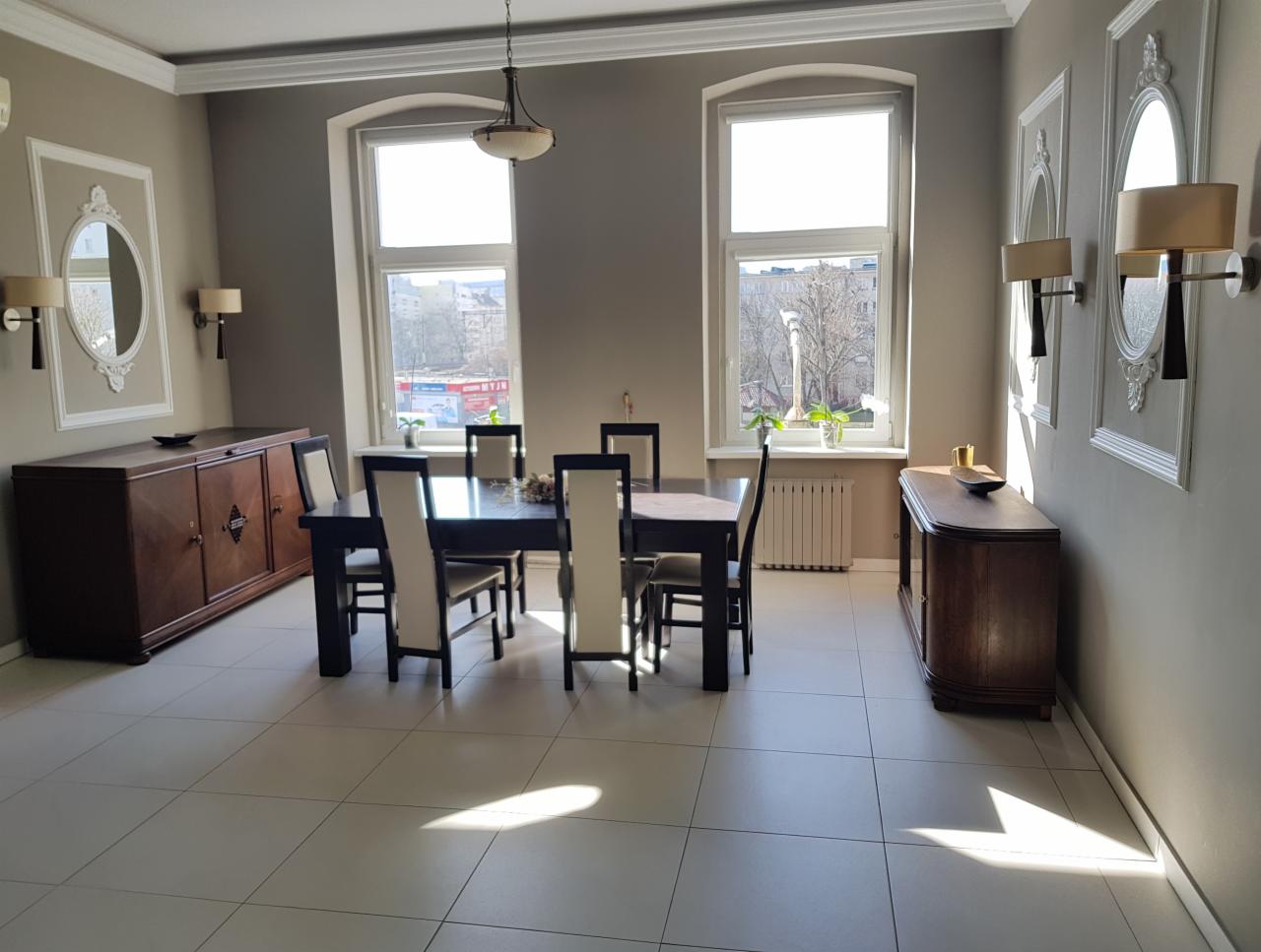 Jasne, przestronne wnętrze jadalni z dużym stołem, krzesłami z kremowymi oparciami, dwiema komodami z ciemnego drewna i lustrami w ozdobnych ramach na ścianach. Dwa duże okna wpuszczają naturalne...