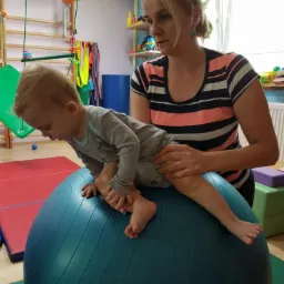 Blondwłosa kobieta w pasiastym topie asekuruje małego chłopca w szarym ubranku podczas ćwiczeń na dużej, niebieskiej piłce gimnastycznej w sali rehabilitacyjnej z drabinkami i półkami.