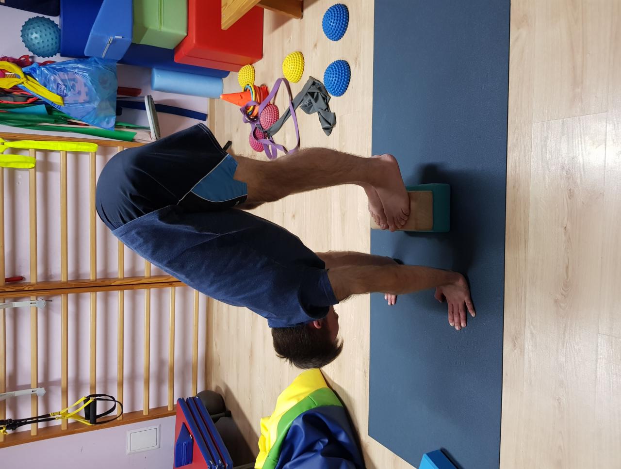 Mężczyzna w pozycji odwróconej na macie do ćwiczeń, stopy oparte na drewnianym bloku, w tle przybory rehabilitacyjne i drabinka.