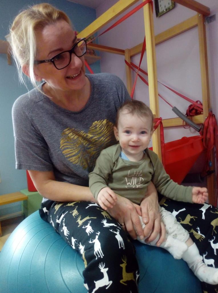 Kobieta w okularach, siedząca na dużej niebieskiej piłce gimnastycznej, trzyma na kolanach uśmiechnięte dziecko w zielonym ubranku, w tle drabinki rehabilitacyjne z czerwonymi gumami.