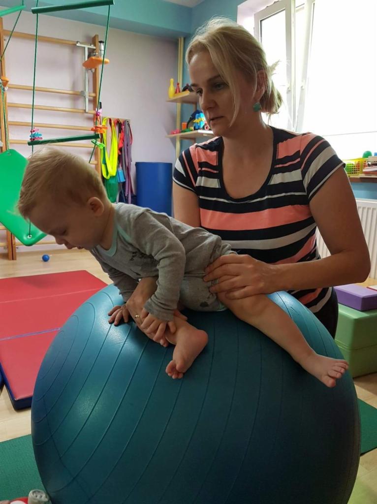 Blondwłosa kobieta w pasiastym topie asekuruje małego chłopca w szarym ubranku podczas ćwiczeń na dużej, niebieskiej piłce gimnastycznej w sali rehabilitacyjnej z drabinkami i półkami.