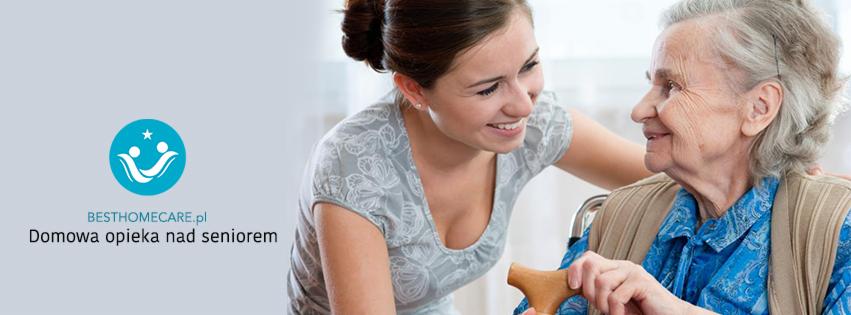 Uśmiechnięta młoda kobieta w bluzce w kwiaty wspiera starszą panią siedzącą na wózku inwalidzkim i trzymającą drewnianą laskę, wzajemny kontakt wzrokowy.