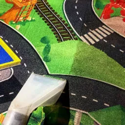 Dysza odkurzacza piorącego na dywanie dziecięcym z motywem ulic, tor&oacute;w kolejowych i zielonej trawy.