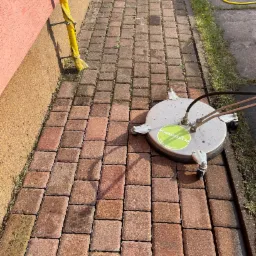 Czyszczenie kostki brukowej przy użyciu profesjonalnej myjki ciśnieniowej z okrągłą głowicą, widoczne różnice w kolorze kostki przed i po czyszczeniu, obok ściana budynku i żółty wąż.