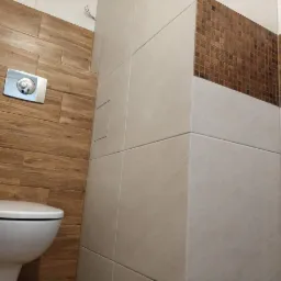 Nowoczesna toaleta z drewnopodobnymi i mozaikowymi płytkami, widoczny przycisk spłukiwania i ukryty zawór rewizyjny.