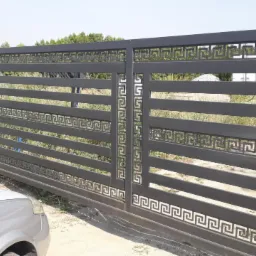 Przesuwna brama wjazdowa z poziomymi panelami i dekoracyjnym greckim wzorem, częściowo zasłonięta przez srebrny samochód, na tle trawy i drzew.