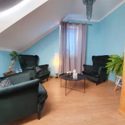 Gabinet z trzema zielonymi fotelami typu uszak, okrągłym stolikiem kawowym z zapalonymi świecami, bonsai i paprotką, oświetlony lampą podłogową i naturalnym światłem z okna dachowego.