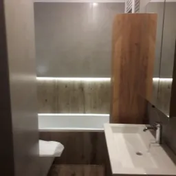 Nowoczesna łazienka z wanną podświetloną listwą LED, umywalką nablatową na drewnianej szafce i toaletą z osłoną, wykończona w szarościach i brązach.