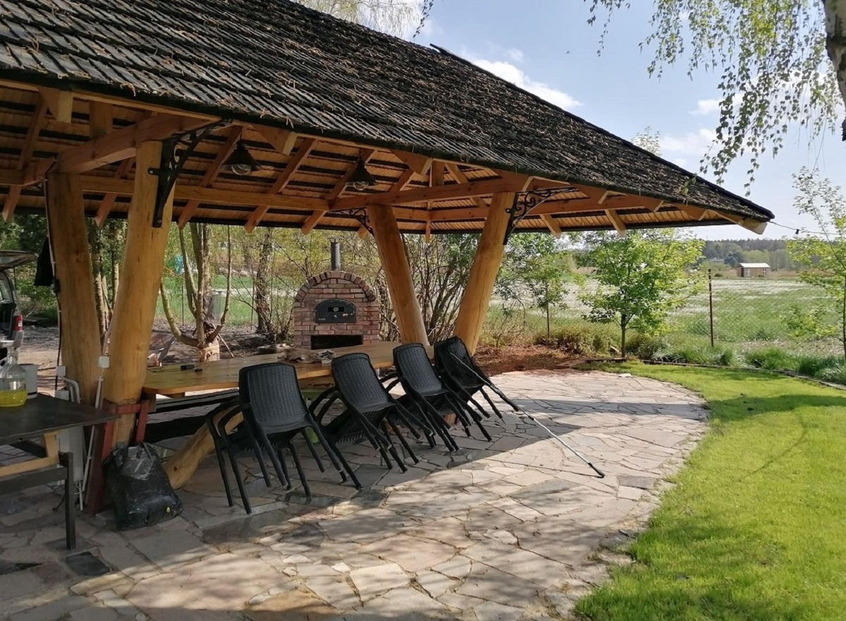 Zadaszona altana z drewnianymi słupami i stołem, piec do pizzy z cegły w tle, taras z nieregularnych płyt kamiennych, czarne krzesła ustawione rzędem.