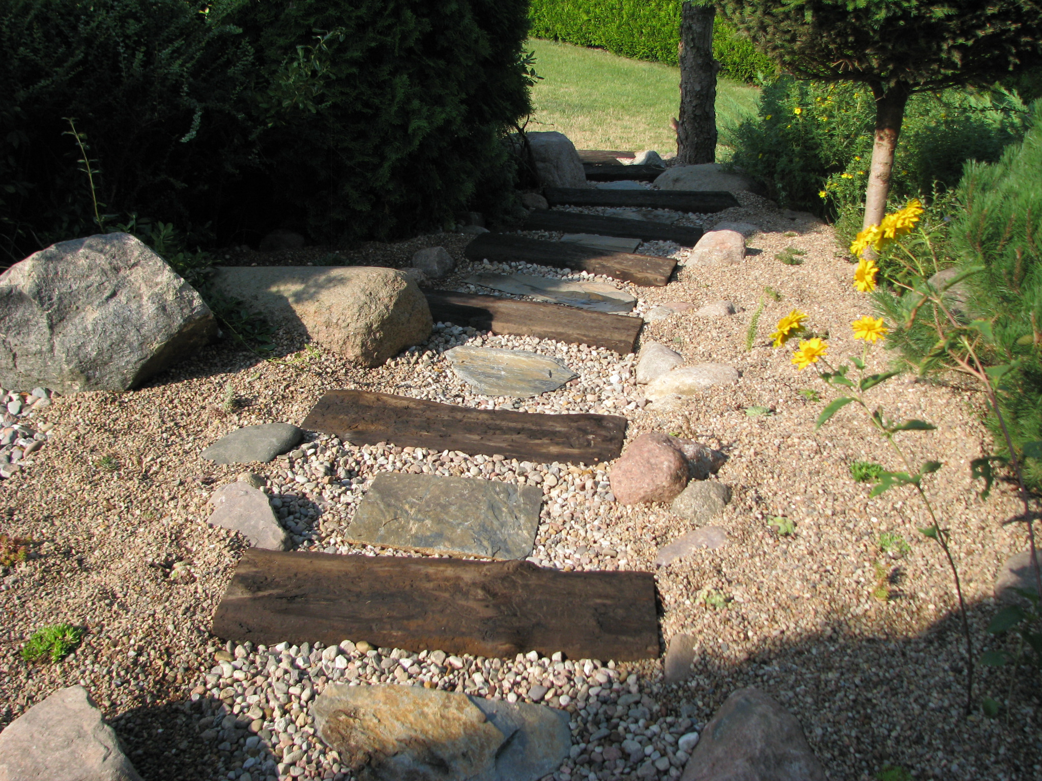 Ścieżka w ogrodzie z drewnianych i kamiennych płyt, wysypana drobnym żwirem, otoczona zielenią i dużymi kamieniami.