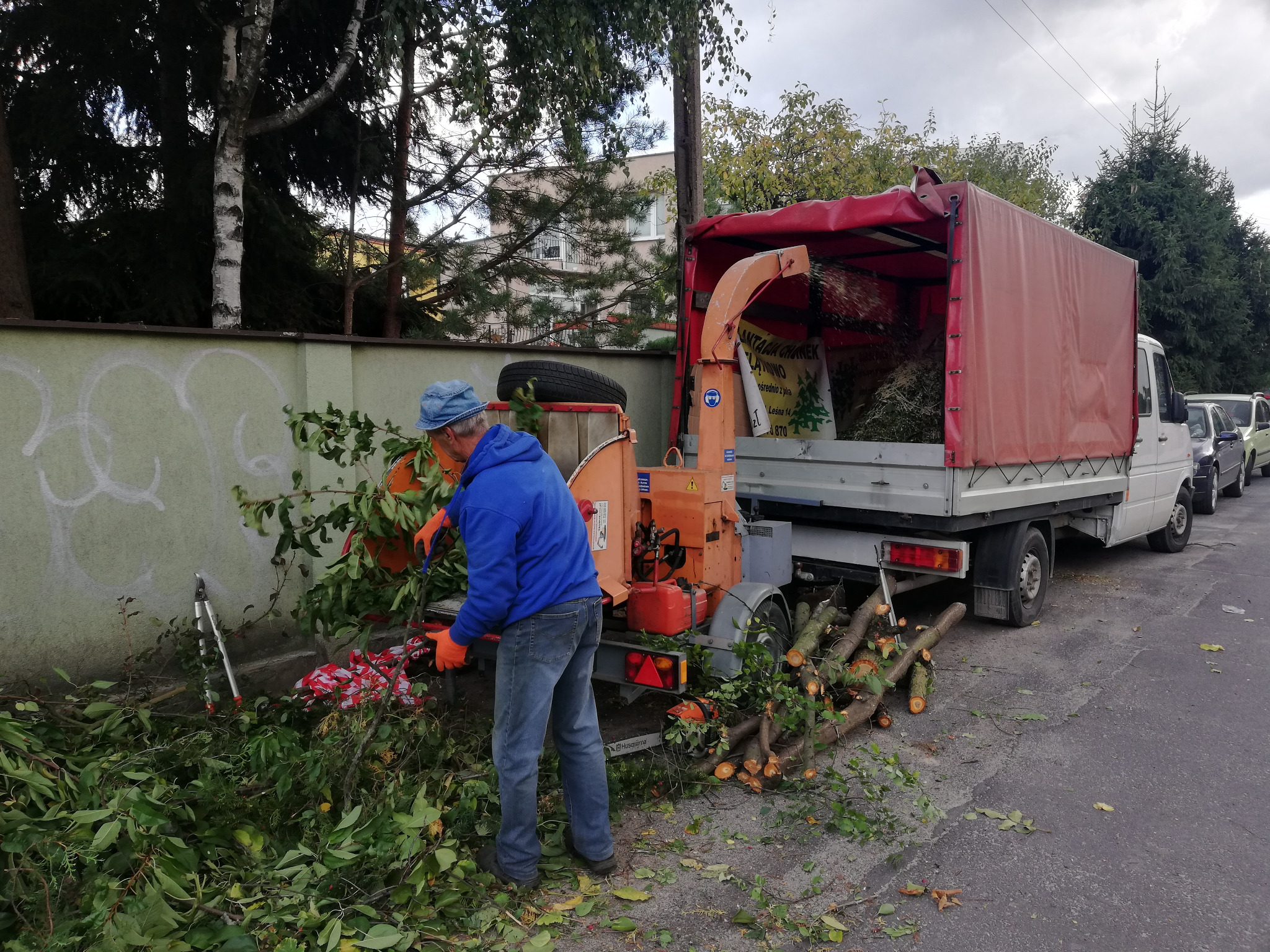Pracownik w niebieskiej bluzie wrzuca gałęzie do rębaka zamontowanego na przyczepie za samochodem dostawczym. Na ziemi leżą pocięte konary i gałęzie, a w tle widać mur i drzewa.