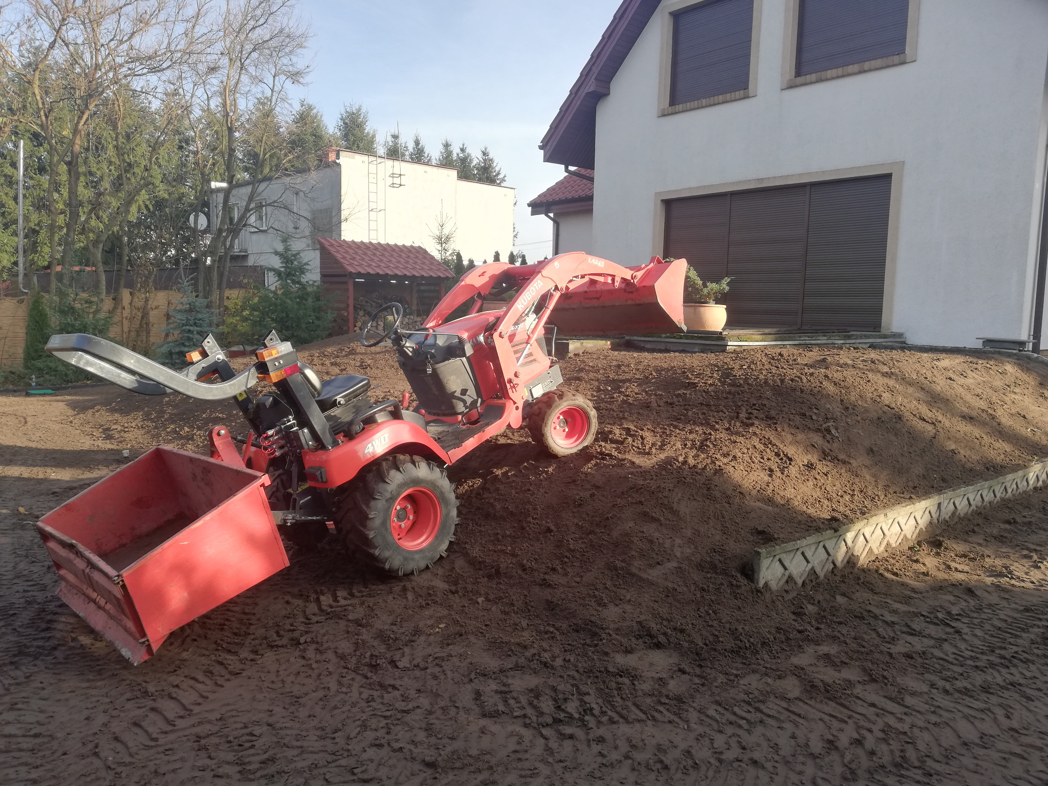 Czerwony mini-traktor Kubota z łyżką i przyczepką na nierównym terenie przed domem z brązowymi roletami.