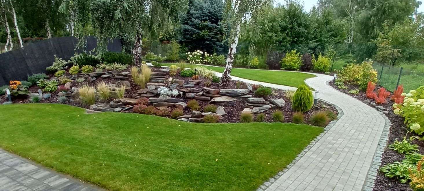 Ogród z malowniczą ścieżką z kostki, skalniakiem z różnorodną roślinnością, drzewami i starannie utrzymanym trawnikiem.