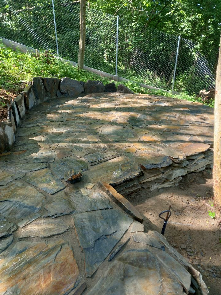 Nierówna ścieżka z nieregularnych, płaskich kamieni w ogrodzie, widoczny fragment metalowego narzędzia wbitego w ziemię obok krawędzi ścieżki.