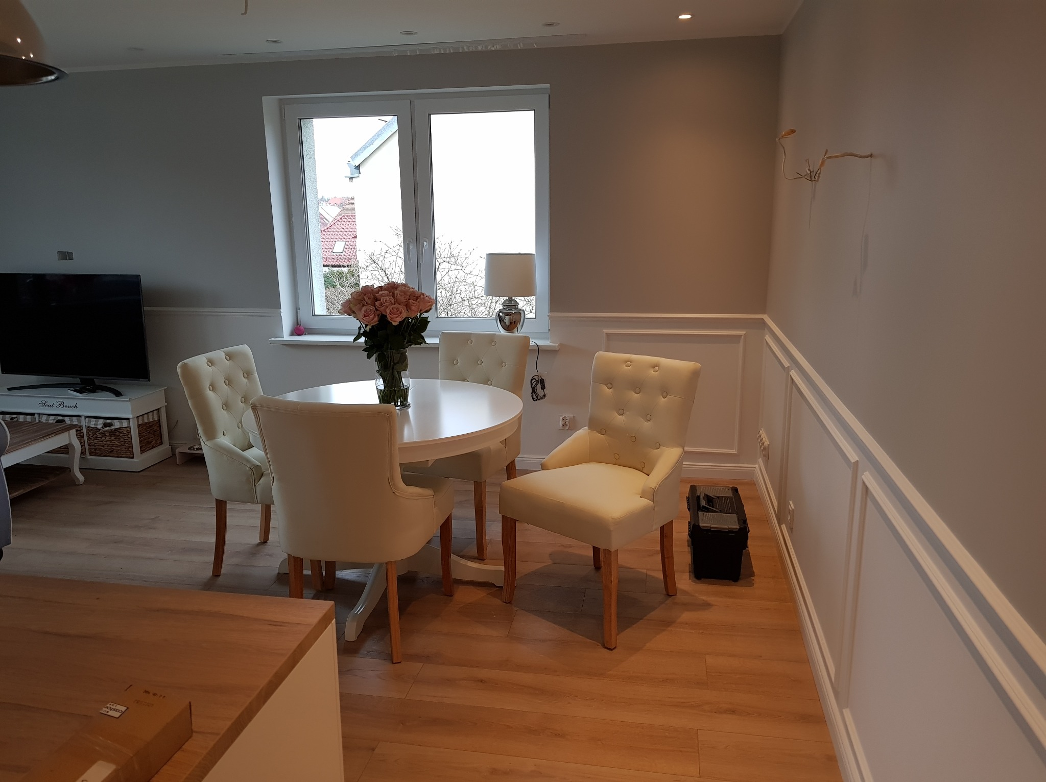 Jasne wnętrze jadalni po remoncie: białe ściany z dekoracyjnymi listwami, okno z widokiem na zewnątrz, okrągły stół z bukietem róż i krzesła w stylu klasycznym.