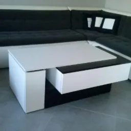 Nowoczesny stolik kawowy w kolorach bieli i czerni z wysuwaną szufladą, dopasowany do narożnej sofy z ciemną tapicerką.
