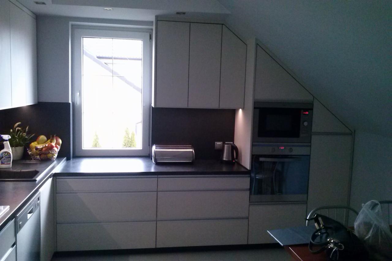 Nowoczesna kuchnia na poddaszu z szarymi frontami, czarnym blatem, oświetleniem LED pod szafkami wiszącymi, zabudowaną mikrofalówką i piekarnikiem, z widokiem na ogród przez okno nad blatem.