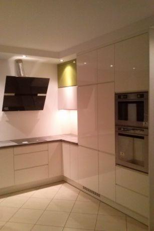 Nowoczesna kuchnia z białymi, lakierowanymi frontami, czarnym okapem i blatem w odcieniach szarości, z piekarnikiem w zabudowie i akcentem w postaci zielonej szafki.