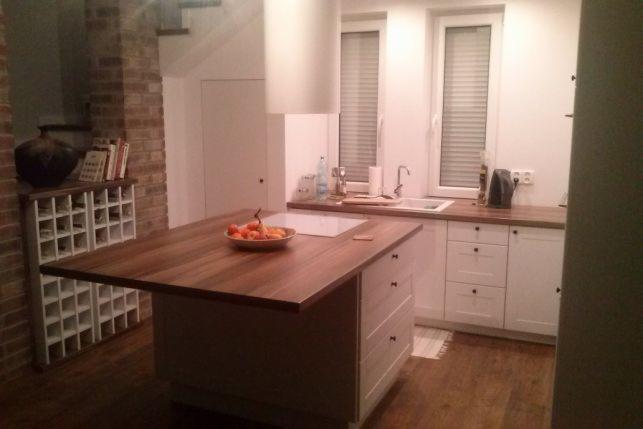 Nowoczesna kuchnia z wyspą z drewnianym blatem, białymi szafkami i płytą indukcyjną, oświetlona światłem wpadającym przez okno z żaluzjami. Na blacie wyspy leżą owoce w misce. W tle widoczny...