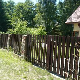 Ogrodzenia Ryn - Ciemnobrązowe, pionowe ogrodzenie z drewnianych sztachet, z kamiennymi słupkami, prowadzące do furtki. W tle zieleń drzew i fragment domu z czerwonym dachem.