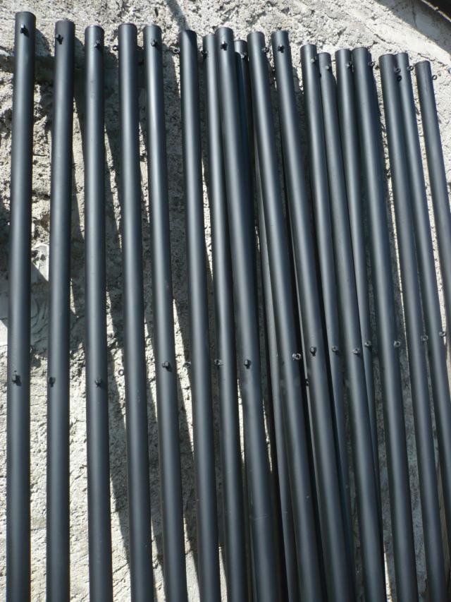 Czarne, metalowe słupki z otworami na śruby, prawdopodobnie elementy konstrukcyjne ogrodzenia, ułożone na szarej, betonowej powierzchni