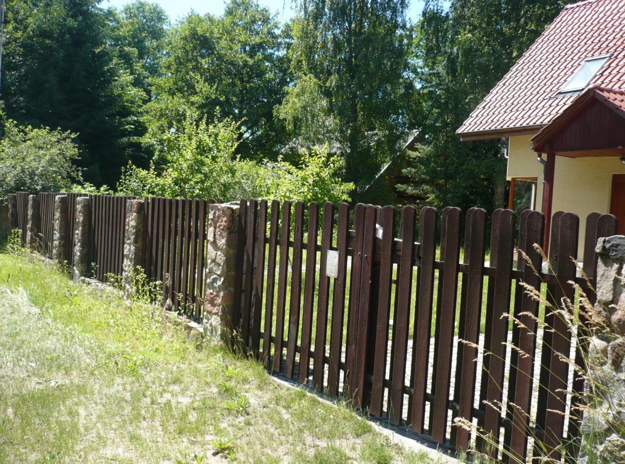 Ciemnobrązowe, pionowe ogrodzenie z drewnianych sztachet, z kamiennymi słupkami, prowadzące do furtki. W tle zieleń drzew i fragment domu z czerwonym dachem.