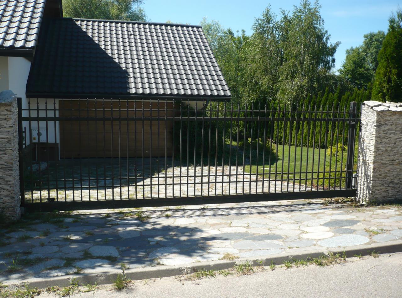 Czarna, przesuwna brama wjazdowa z metalowymi prętami, osadzona na kamiennych słupkach, na tle domu z garażem i zadbanego trawnika.