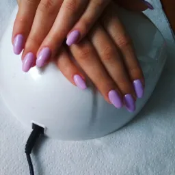 Dłonie z świeżo pomalowanymi, owalnymi paznokciami w pastelowym odcieniu fioletu, suszone w lampie UV na tle białej, teksturowanej powierzchni.