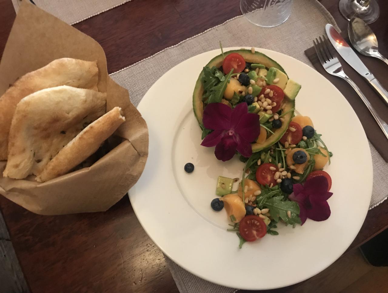 Melonowa misa wypełniona sałatką z rukoli, pomidorków cherry, awokado, borówek i orzeszków piniowych, ozdobiona fioletowymi storczykami, obok koszyk z pieczywem.