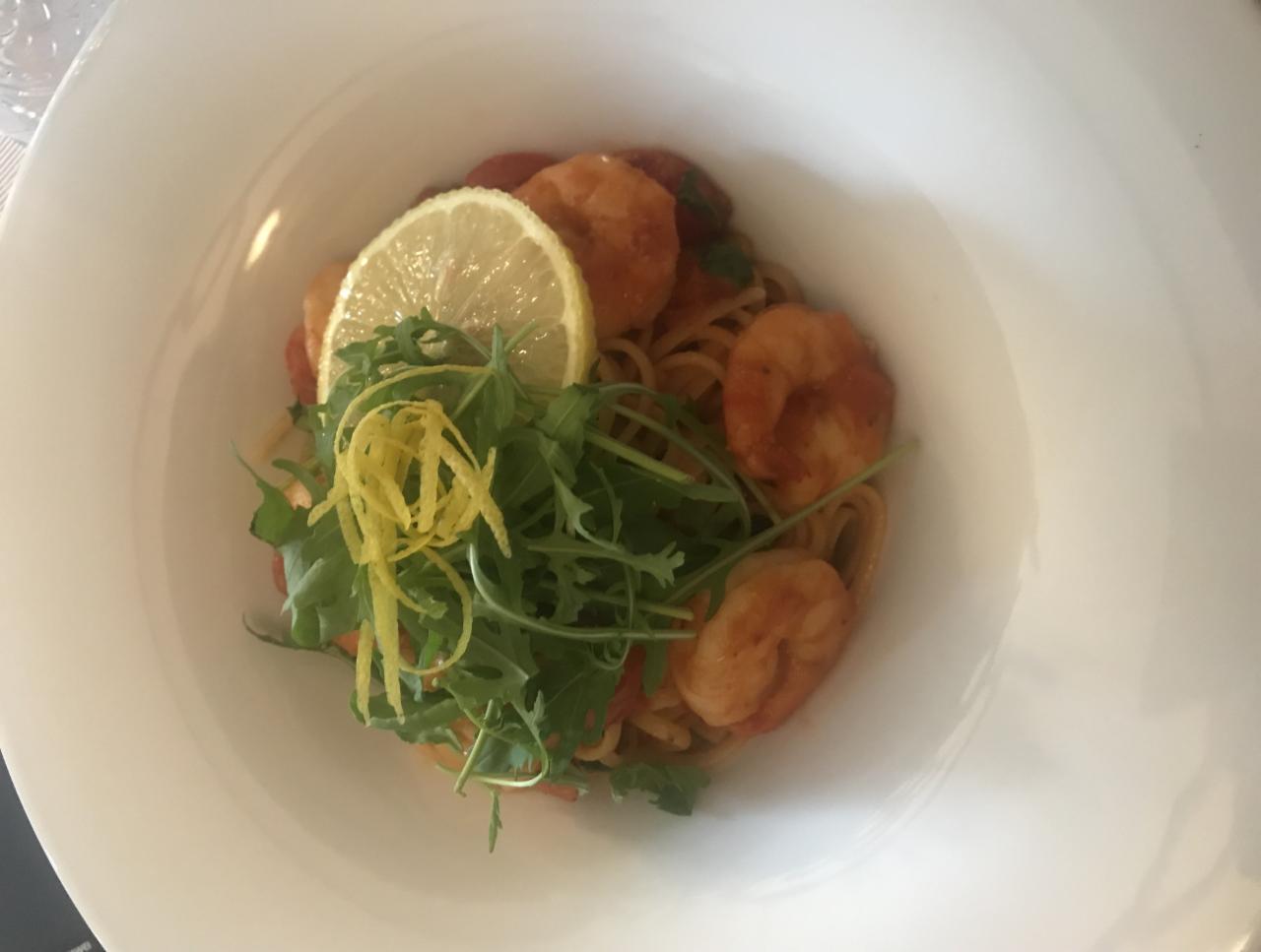 Porcja spaghetti z krewetkami, pomidorami, rukolą i dekoracją z cytryny w białym, głębokim talerzu.