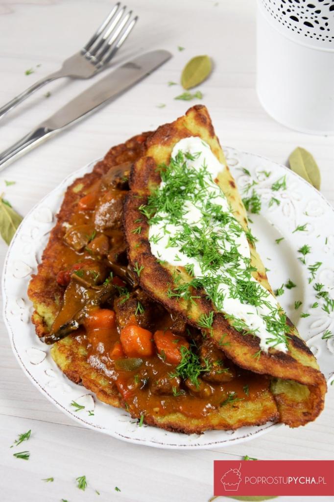 Placki ziemniaczane z gulaszem warzywnym i kleksem śmietany, posypane świeżym koperkiem, podane na białym, zdobionym talerzu, ze sztućcami i dekoracyjnymi listkami laurowymi w tle.