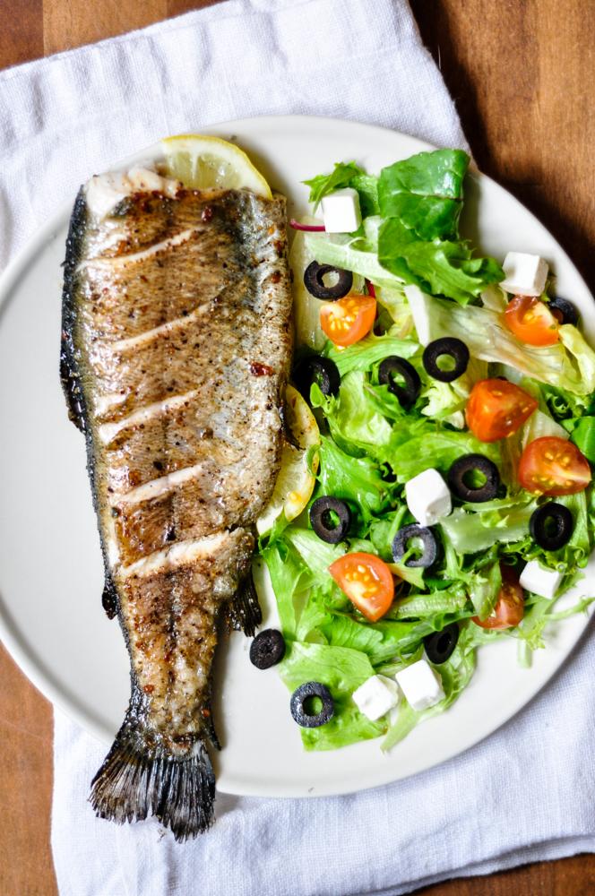 Grilowany pstrąg z plasterkiem cytryny, podany z sałatką ze świeżych warzyw, czarnych oliwek i kostek sera feta na białym talerzu.