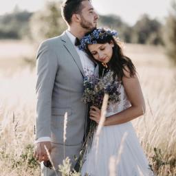 Perspective fotografia - Para młoda w plenerze: panna młoda w białej sukni z koronkową górą i wiankiem z chabrów, opiera głowę na ramieniu pana młodego w szarym garniturze z muchą, trzyma bukiet polnych kwiatów, wysoka...