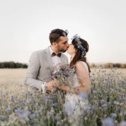 Pocałunek pary młodej w plenerze, otoczonej polem kwitnących chabrów, pan młody w szarym garniturze z muszką, panna młoda w wianku z kwiatów.
