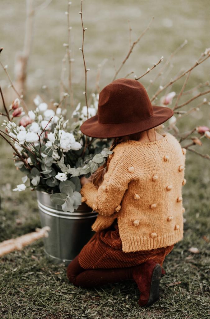 Dziewczynka w brązowym kapeluszu i musztardowym swetrze z pomponami, klęcząca obok metalowego wiadra z białymi kwiatami i gałązkami, widok z tyłu, naturalne światło.