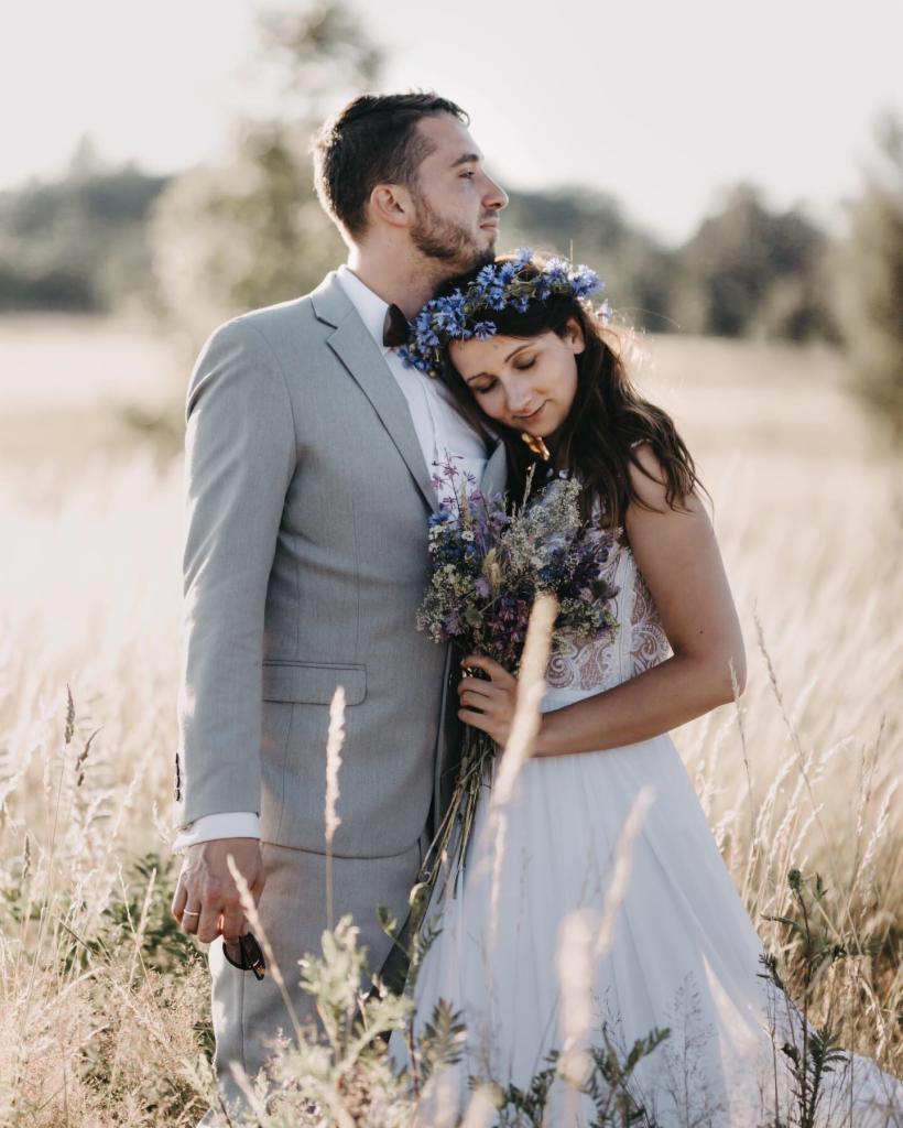 Para młoda w plenerze: panna młoda w białej sukni z koronkową górą i wiankiem z chabrów, opiera głowę na ramieniu pana młodego w szarym garniturze z muchą, trzyma bukiet polnych kwiatów, wysoka...