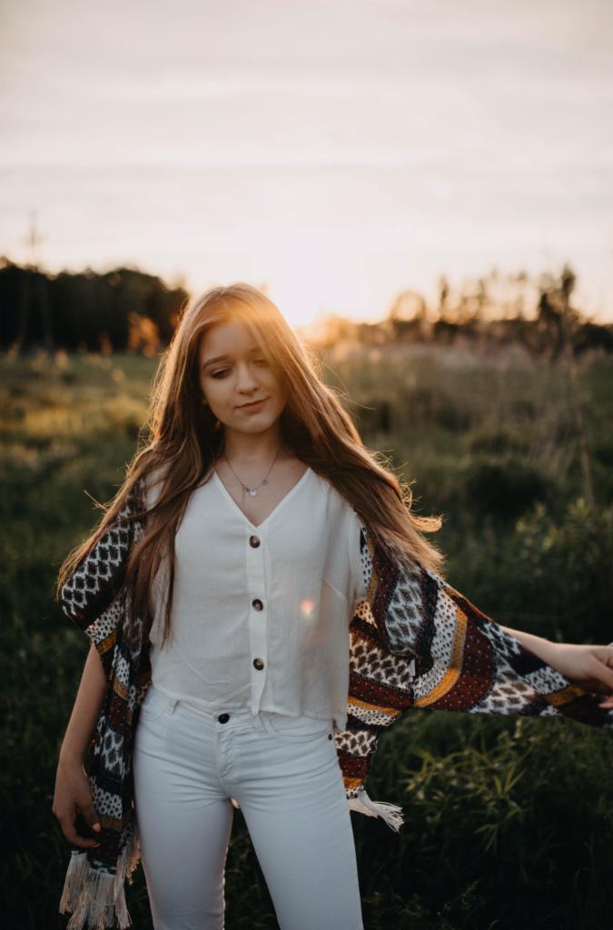 Młoda kobieta w białym stroju i wzorzystym szalu pozuje na łące w ciepłym świetle zachodzącego słońca.