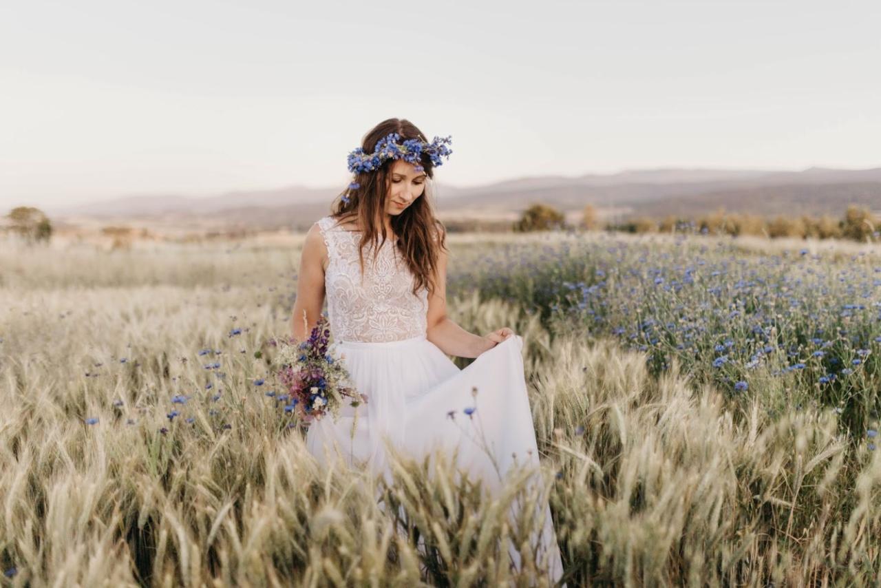 Panna młoda w koronie z błękitnych kwiatów i bukietem polnych kwiatów, spacerująca po polu pszenicy i bławatków w delikatnym świetle zachodzącego słońca.