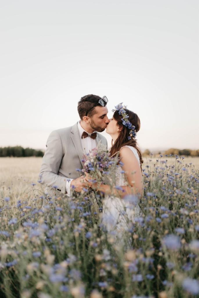 Pocałunek pary młodej w plenerze, otoczonej polem kwitnących chabrów, pan młody w szarym garniturze z muszką, panna młoda w wianku z kwiatów.