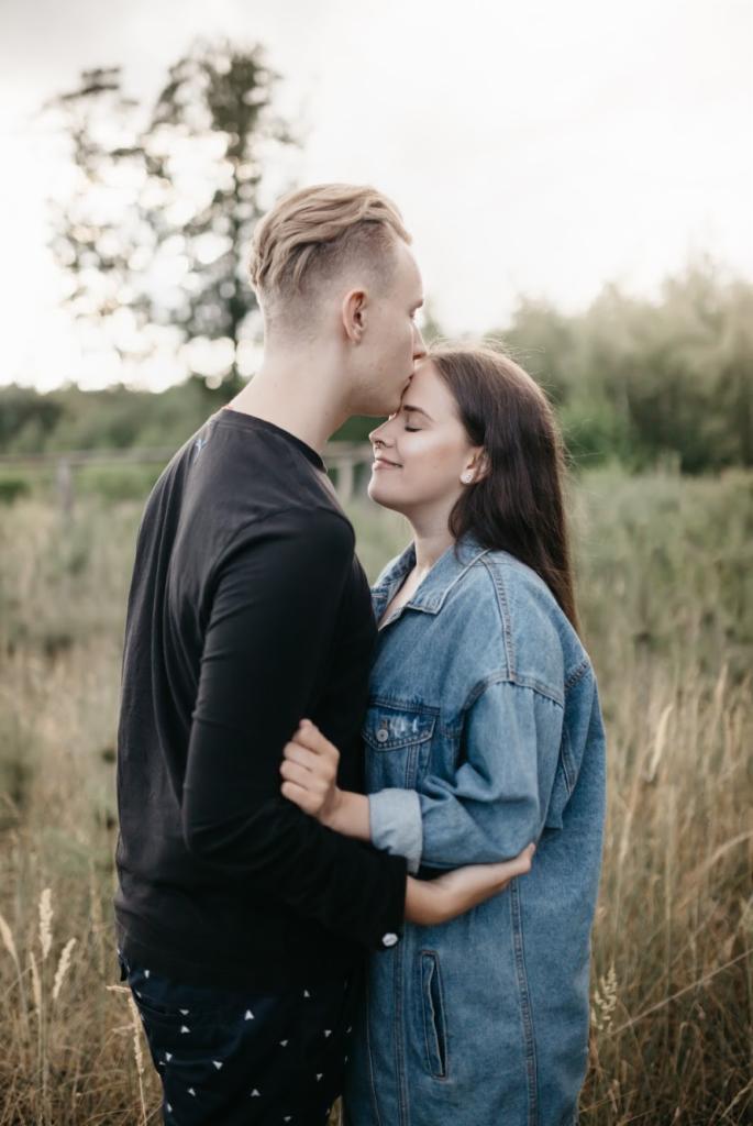 Młoda para w objęciach na tle łąki, mężczyzna całuje kobietę w czoło, ona ma zamknięte oczy i się uśmiecha.