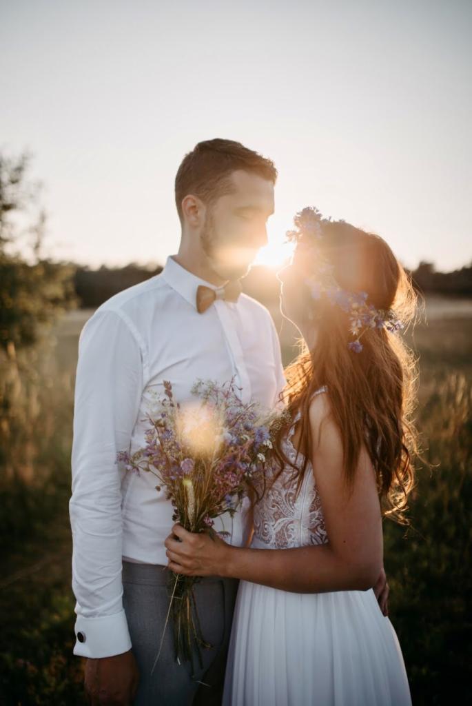 Para młoda w plenerze o zachodzie słońca, panna młoda w wianku z polnych kwiatów i sukni z koronkową górą trzyma bukiet, pan młody w białej koszuli i drewnianej muszce.