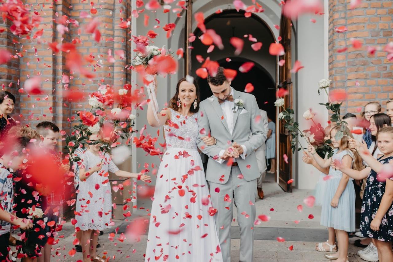 Para młoda wychodzi z kościoła, obsypywana czerwonymi płatkami róż przez dzieci, panna młoda trzyma bukiet białych róż.