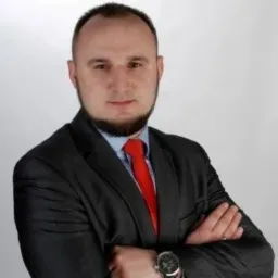 Mężczyzna w garniturze z czerwoną kravatą, skrzyżowane ręce, zegarek na nadgarstku, broda, krótko ostrzyżone włosy. Portret biznesowy na jasnym tle.