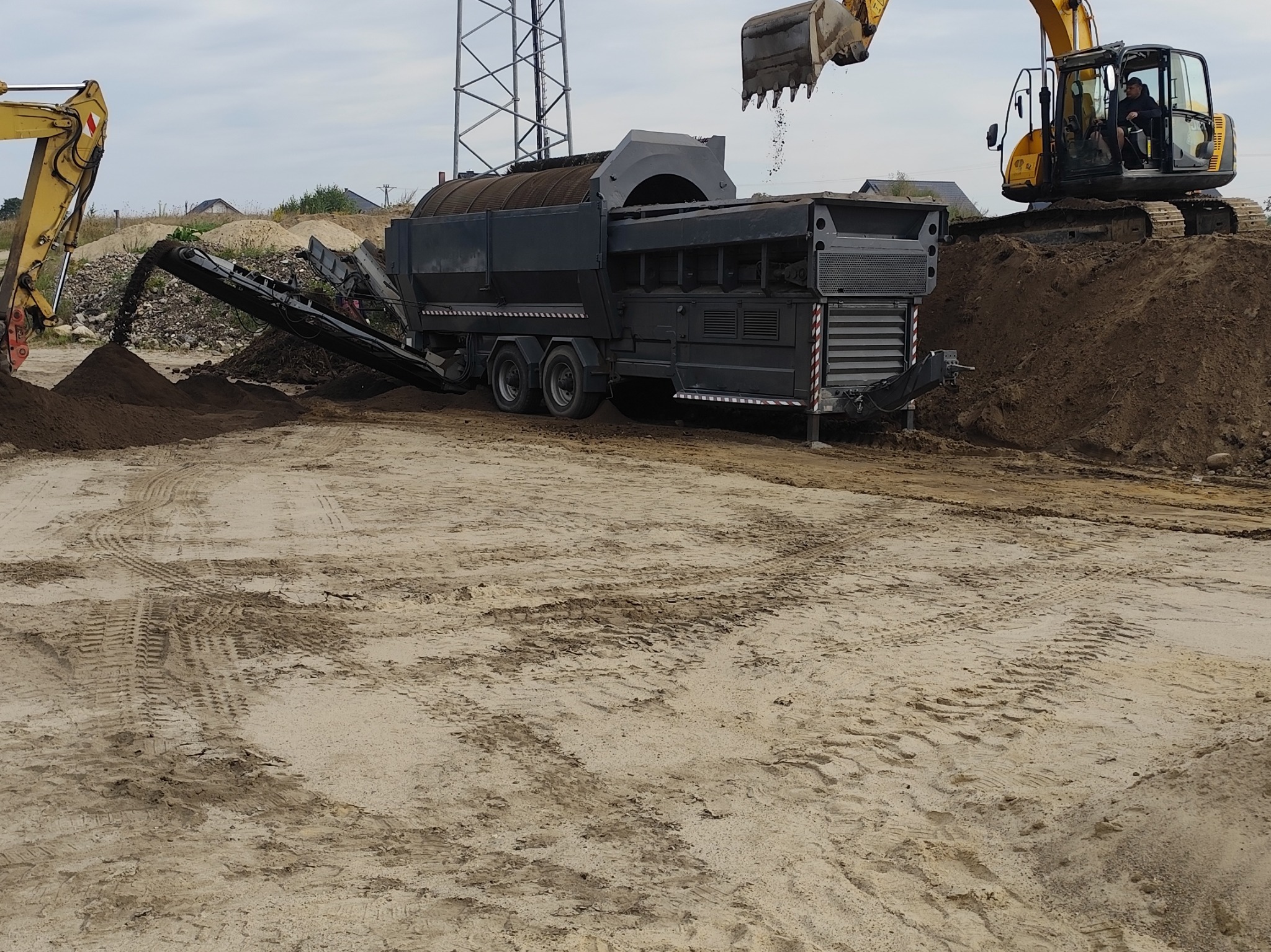 Mobilna maszyna do przesiewania i kruszenia gruzu na placu budowy, z dwoma koparkami w tle, jedna z operatorem w kabinie, druga z łyżką w trakcie załadunku materiału do przesiewacza.