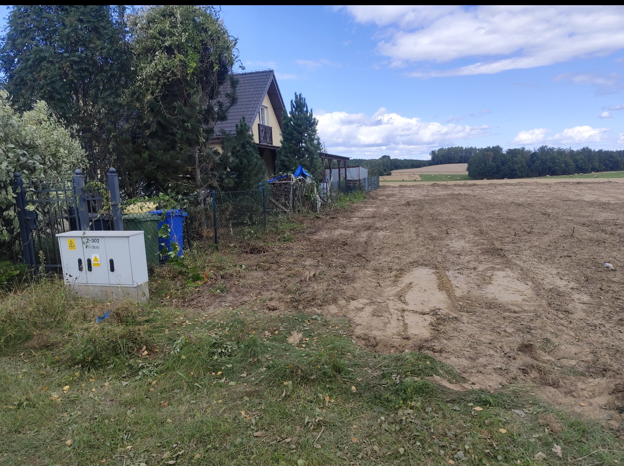 Teren po wycince drzew z widocznym domem w tle. Świeżo uprzątnięta działka z pozostałościami po pracach ziemnych i zielenią. Błękitne niebo z chmurami.