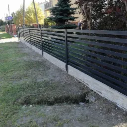 Nowoczesne, poziome, metalowe ogrodzenie w kolorze grafitowym, osadzone na betonowej podmurówce, z widocznym wykopem wzdłuż ogrodzenia i osobą w tle.