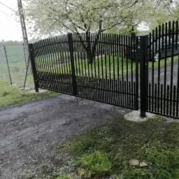 Czarna, metalowa brama wjazdowa z pionowymi prętami, otwierana na posesję z widocznym samochodem w tle, na nieutwardzonej drodze dojazdowej.