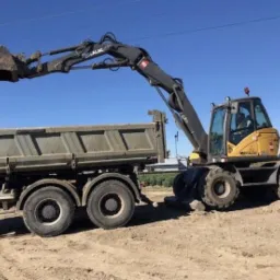 Żółto-szara koparka kołowa Mecalac 714MW załadowuje ziemię do skrzyni wywrotki na tle błękitnego nieba.