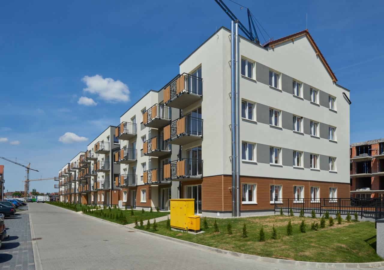 Nowoczesny budynek mieszkalny z balkonami, wykończony drewnianą okładziną, z widoczną instalacją odprowadzającą spaliny i żółtą skrzynką gazową na tle błękitnego nieba i żurawi budowlanych.
